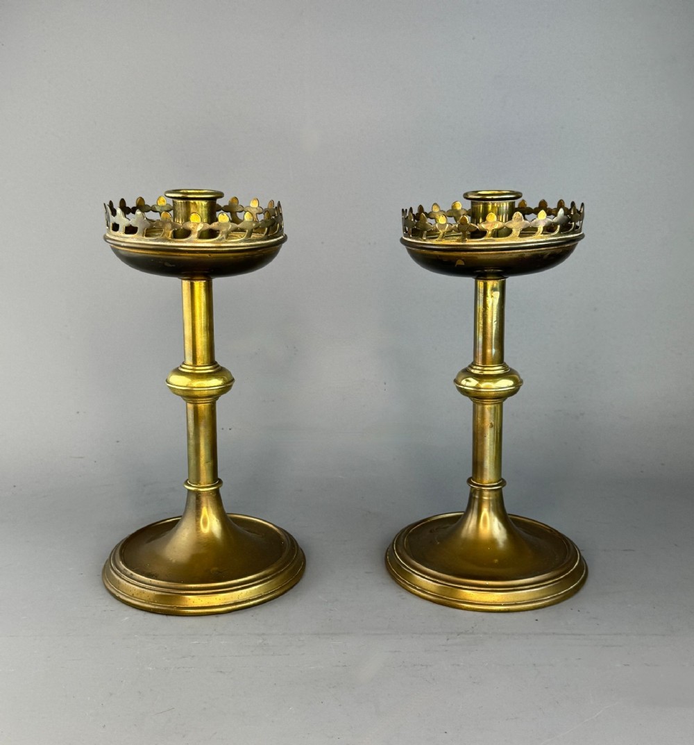 A pair of Victorian Gothic Revival brass candlesticks featuring turned shafts, circular bases, and pierced crown drip pans.