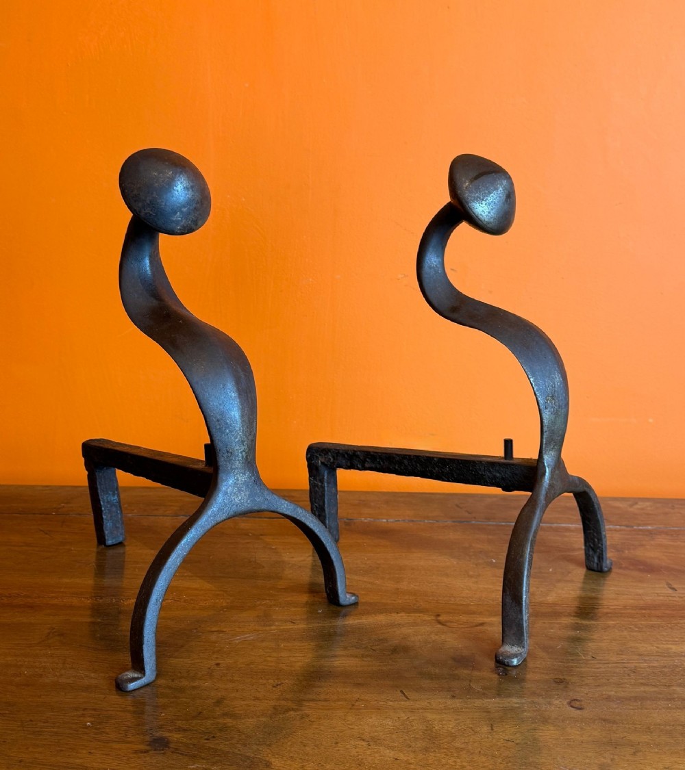 A pair of hand-forged dark wrought iron Arts and Crafts style andirons featuring curved upright stems with rounded disc finials.