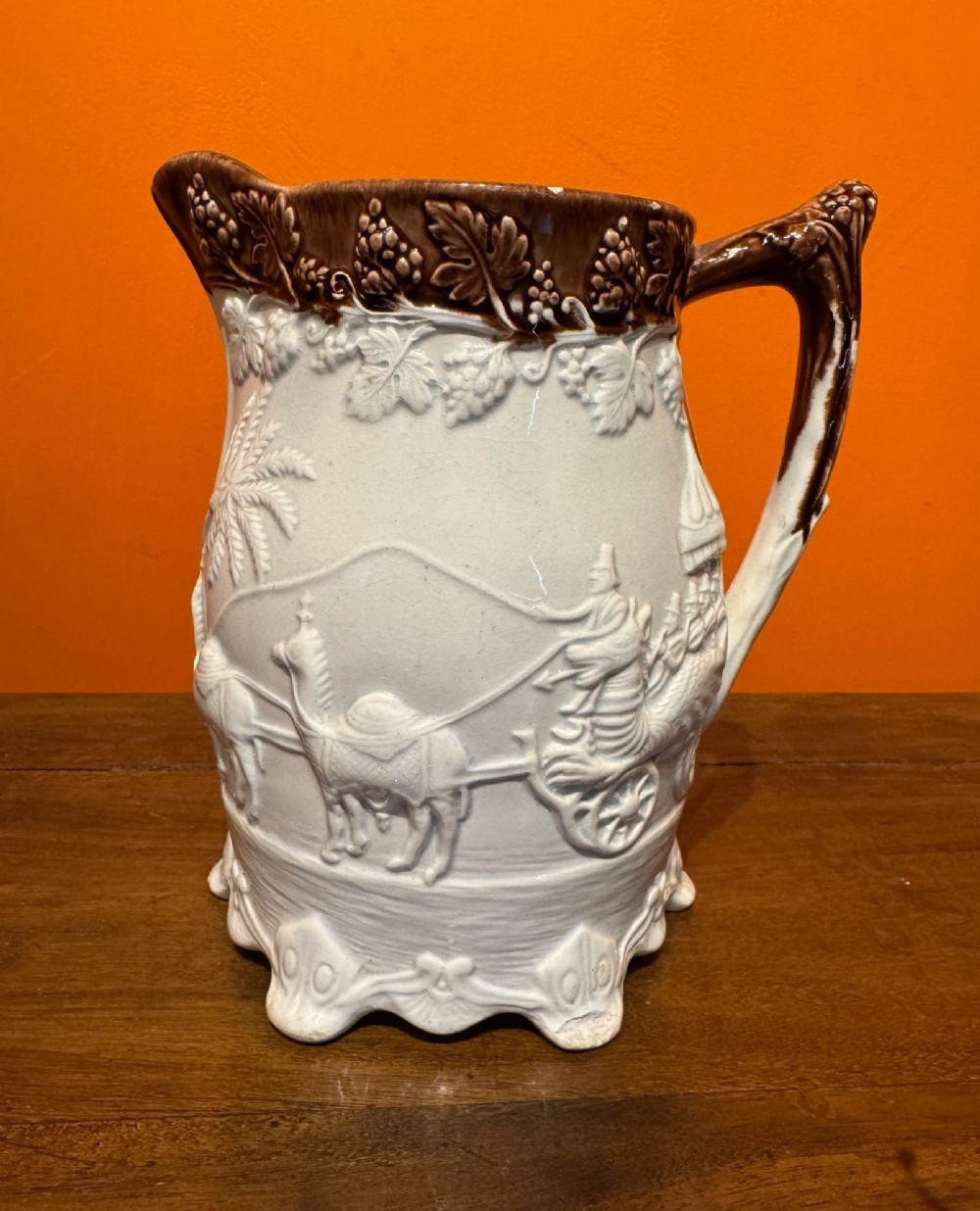 A Victorian relief-moulded earthenware jug featuring an embossed Indian carriage scene on a cream body with a brown glazed grapevine rim and handle.