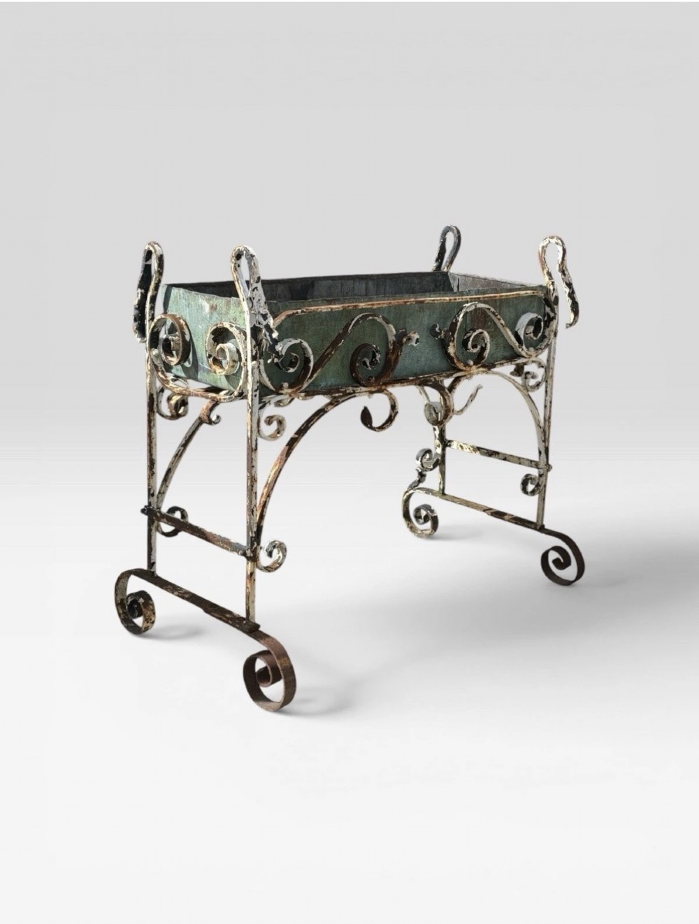 A white painted wrought iron planter stand featuring decorative scrollwork legs holding a rectangular green oxidised metal trough liner. View II