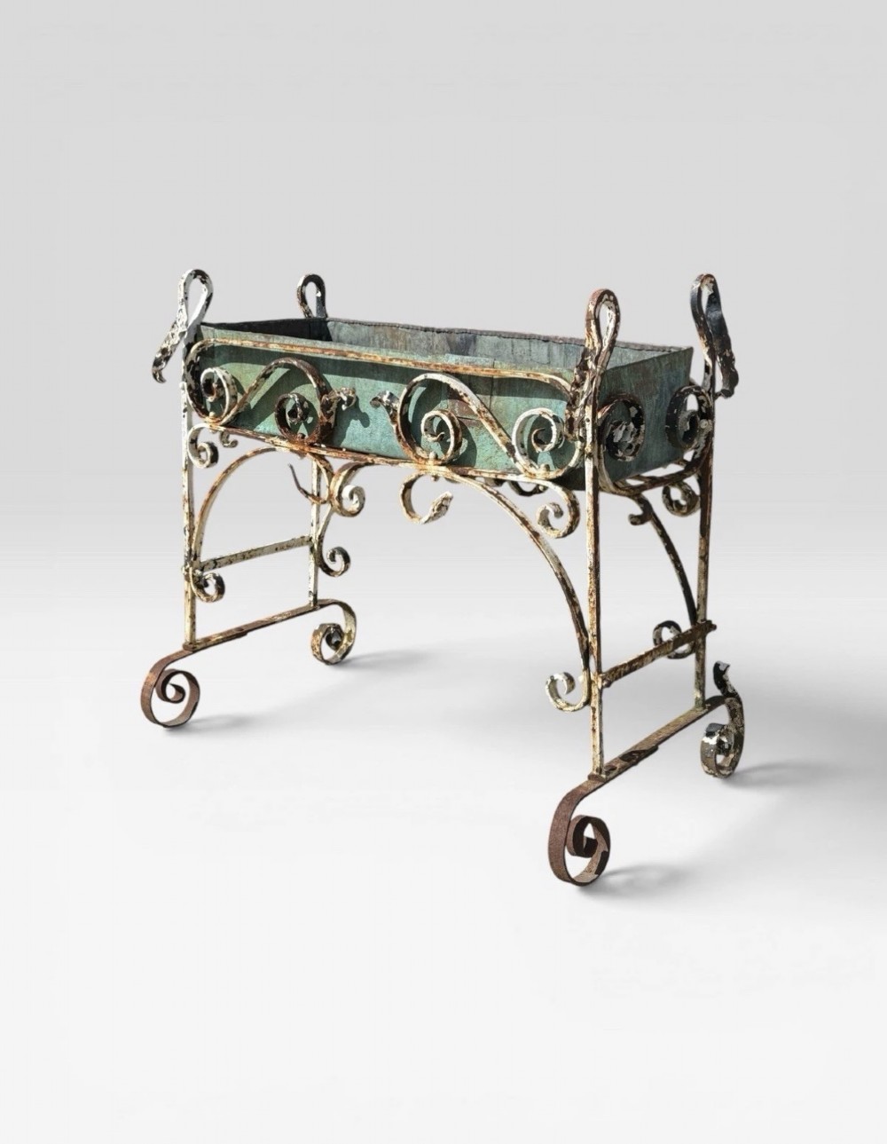 A white painted wrought iron planter stand featuring decorative scrollwork legs holding a rectangular green oxidised metal trough liner. View III