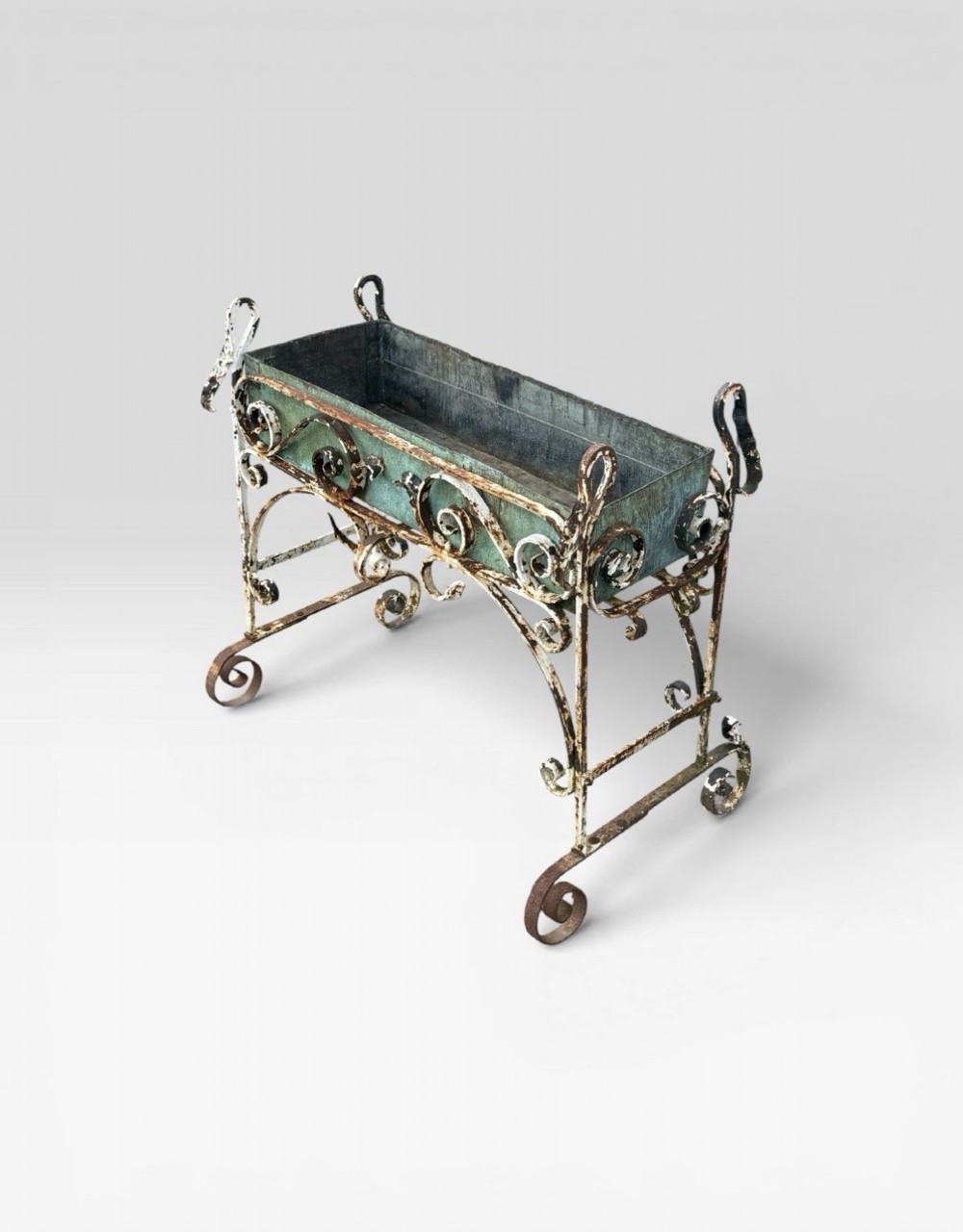 A white painted wrought iron planter stand featuring decorative scrollwork legs holding a rectangular green oxidised metal trough liner. View VII