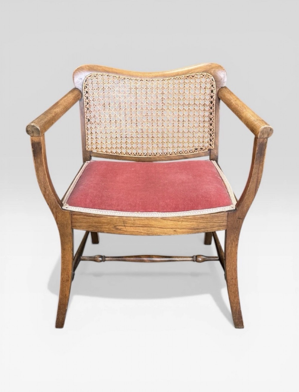 An antique childs wooden armchair featuring a cane woven backrest, swept armrests, turned stretchers, and a red velvet upholstered drop-in seat.