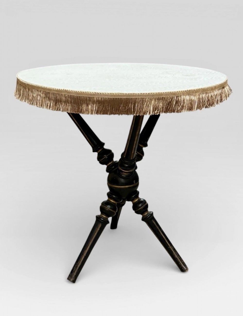 A Victorian Aesthetic Movement ebonised gypsy table with turned tripod legs, gilt highlights, and a cream fabric top with metallic fringe.