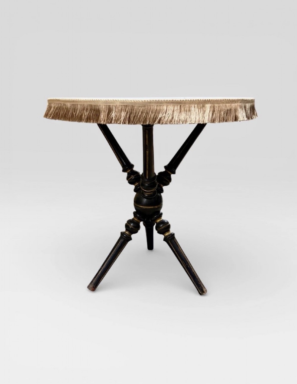 A Victorian Aesthetic Movement ebonised gypsy table with turned tripod legs, gilt highlights, and a cream fabric top with metallic fringe. View VI