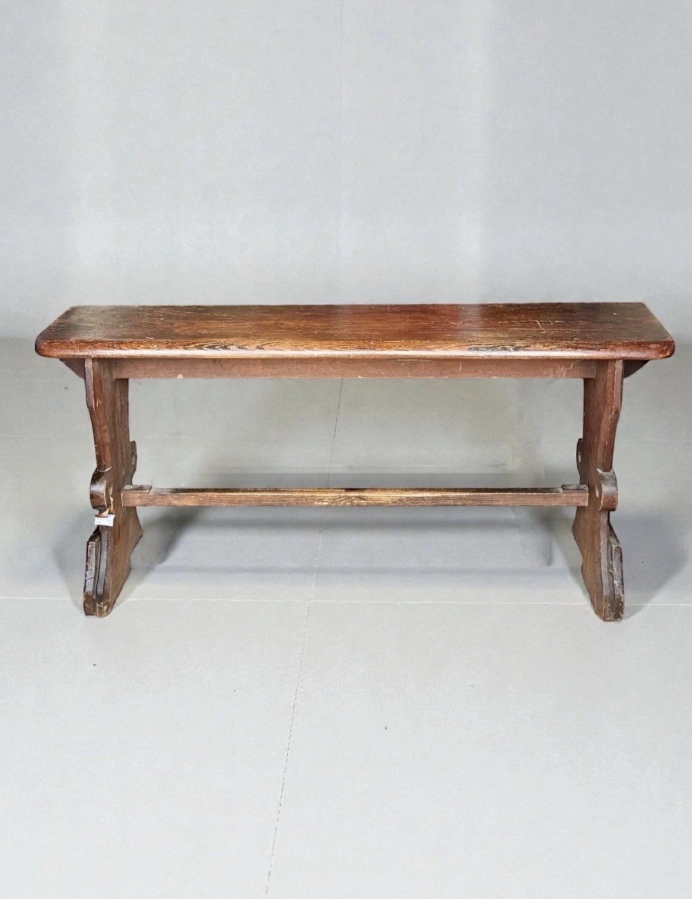 A dark warm brown antique pine organist bench with shaped side trestles and a low central connecting stretcher.