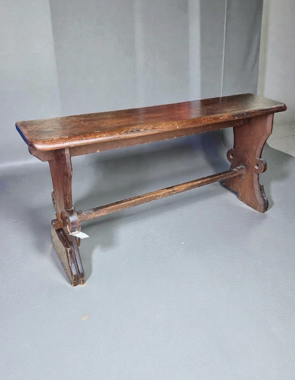 A dark warm brown antique pine organist bench with shaped side trestles and a low central connecting stretcher. View II