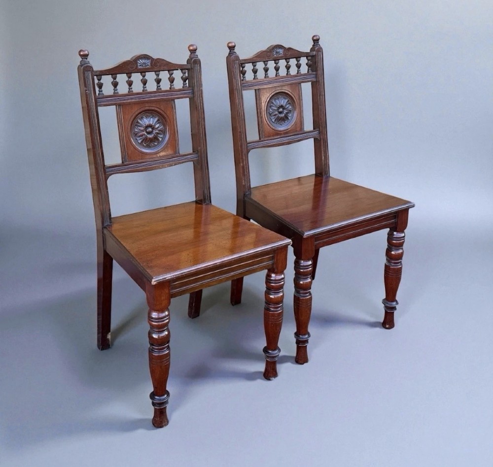 A pair of Victorian mahogany hall chairs featuring carved rosette back panels, spindled top rails, solid wood seats, and turned front legs. View II