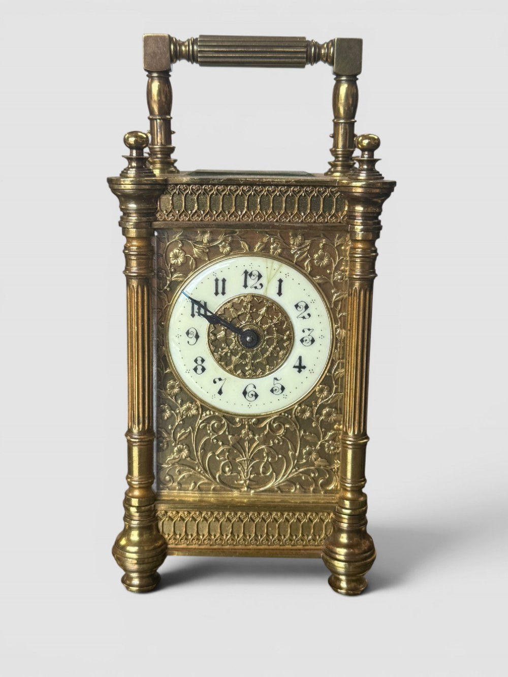 An ornate brass carriage clock featuring fluted corner columns, top finials, a carrying handle, and an engraved floral faceplate surrounding a circular enamel dial.