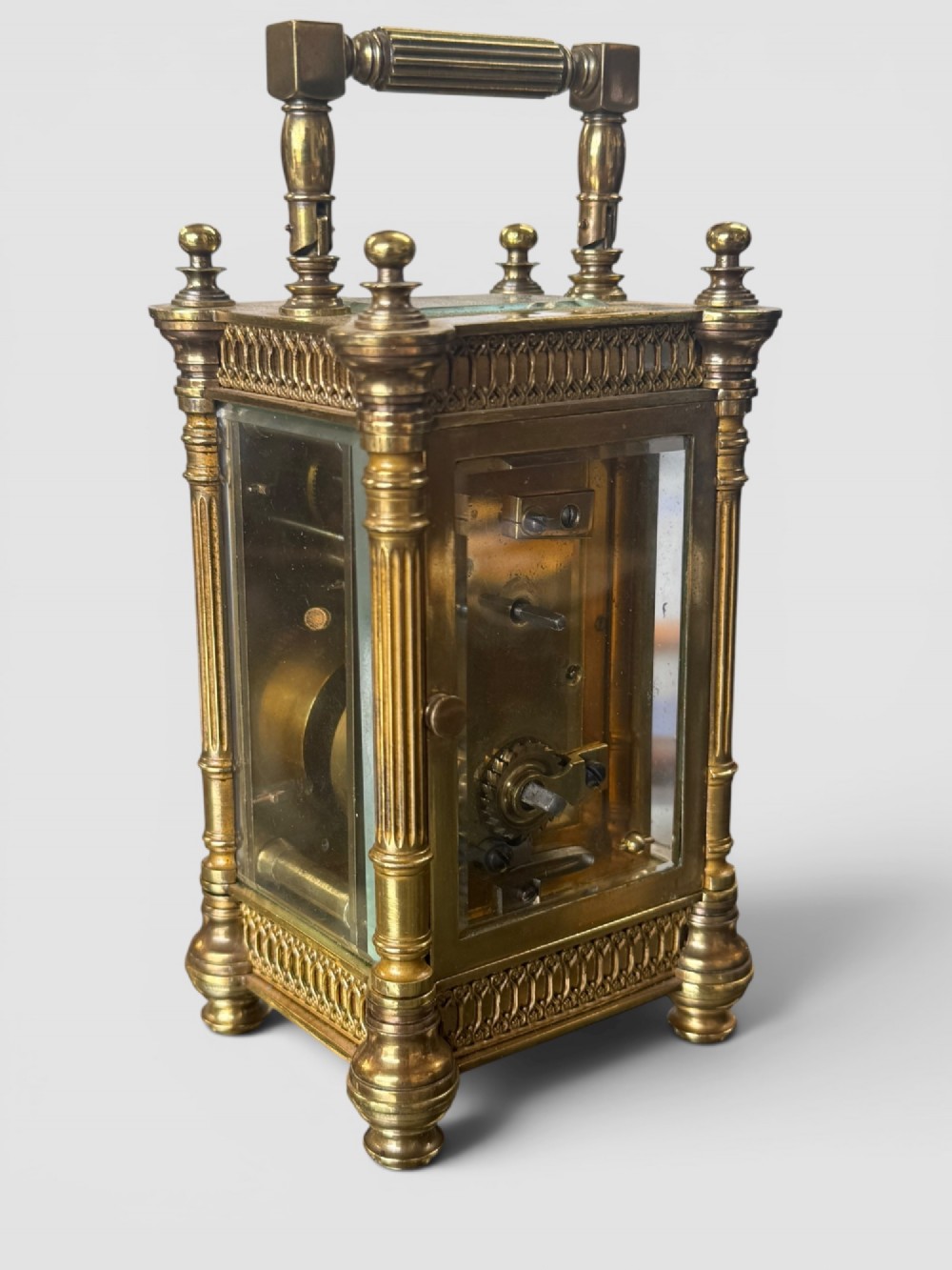 An ornate brass carriage clock featuring fluted corner columns, top finials, a carrying handle, and an engraved floral faceplate surrounding a circular enamel dial. View IV