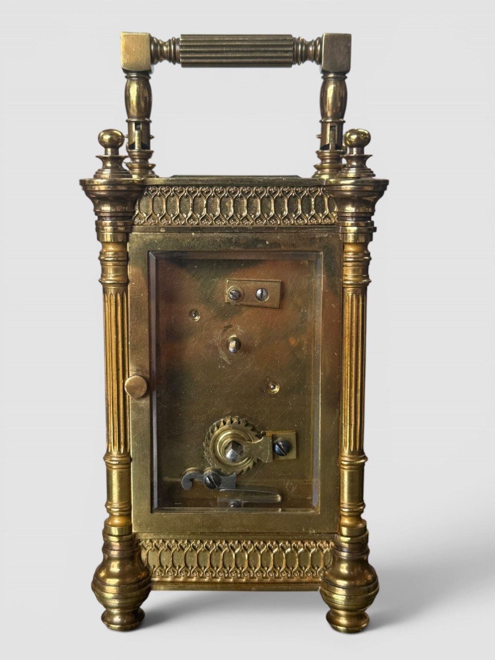 An ornate brass carriage clock featuring fluted corner columns, top finials, a carrying handle, and an engraved floral faceplate surrounding a circular enamel dial. View V