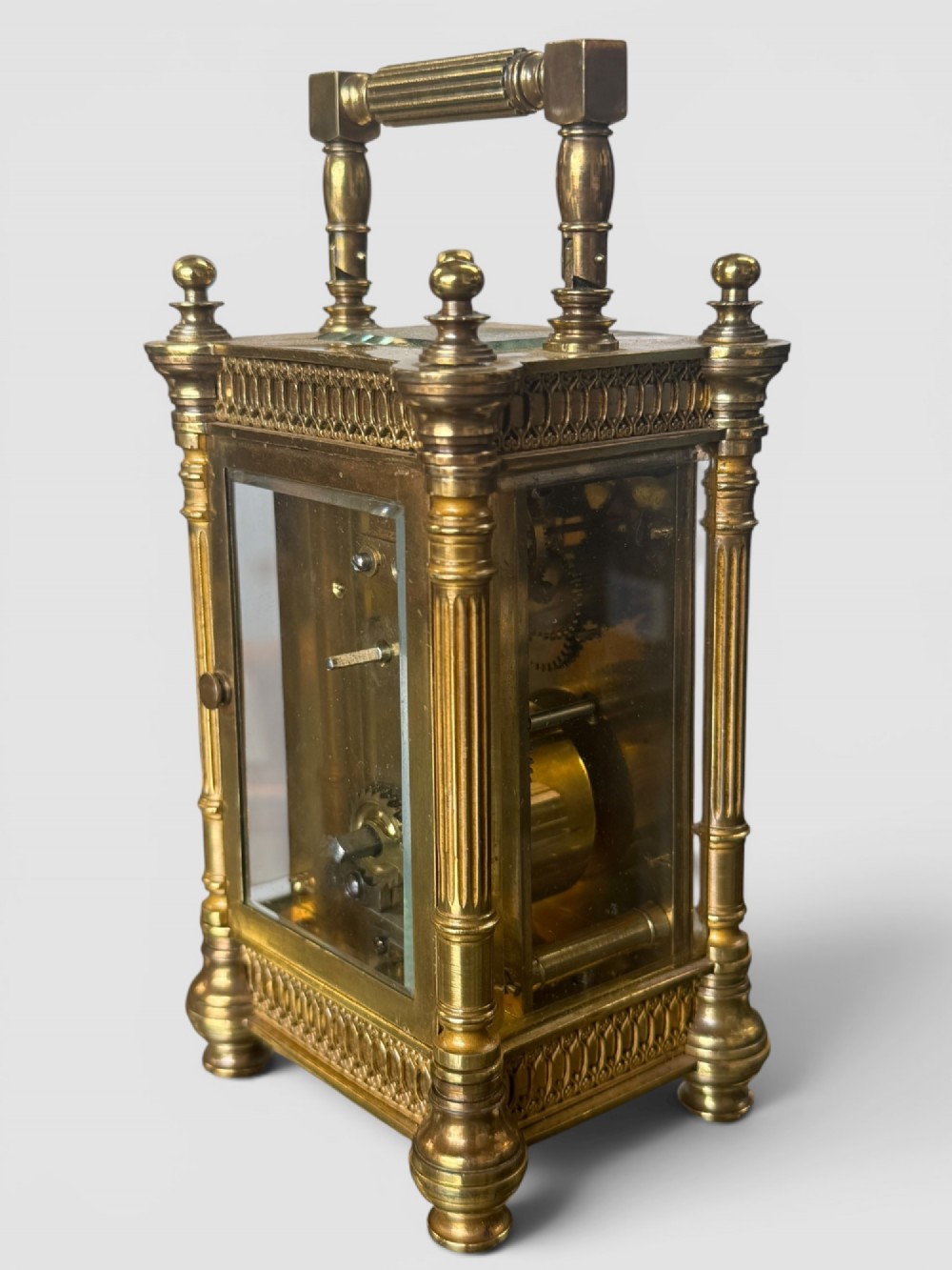 An ornate brass carriage clock featuring fluted corner columns, top finials, a carrying handle, and an engraved floral faceplate surrounding a circular enamel dial. View VI