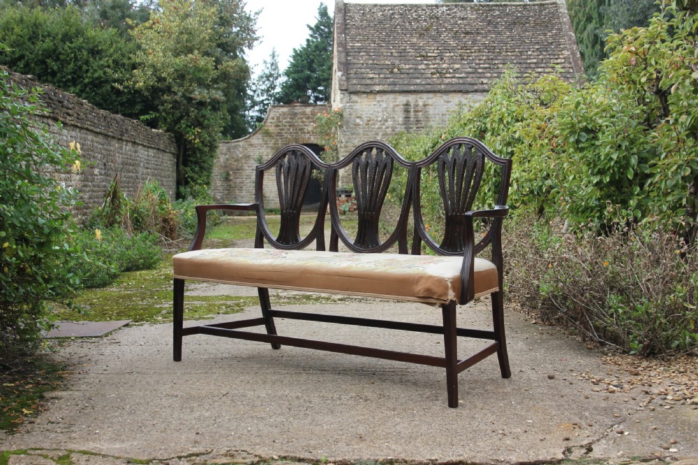 A 19th century mahogany triple shield back settee with carved splats, curved arms, and an upholstered seat cushion.