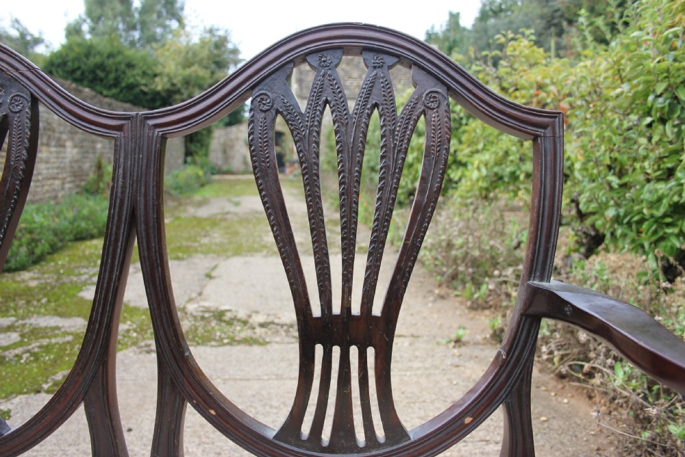 A 19th century mahogany triple shield back settee with carved splats, curved arms, and an upholstered seat cushion. View V