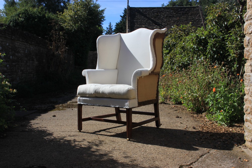An 18th-century wingback armchair upholstered in off-white calico lining with dark mahogany legs, an H-stretcher frame, and brass castors.