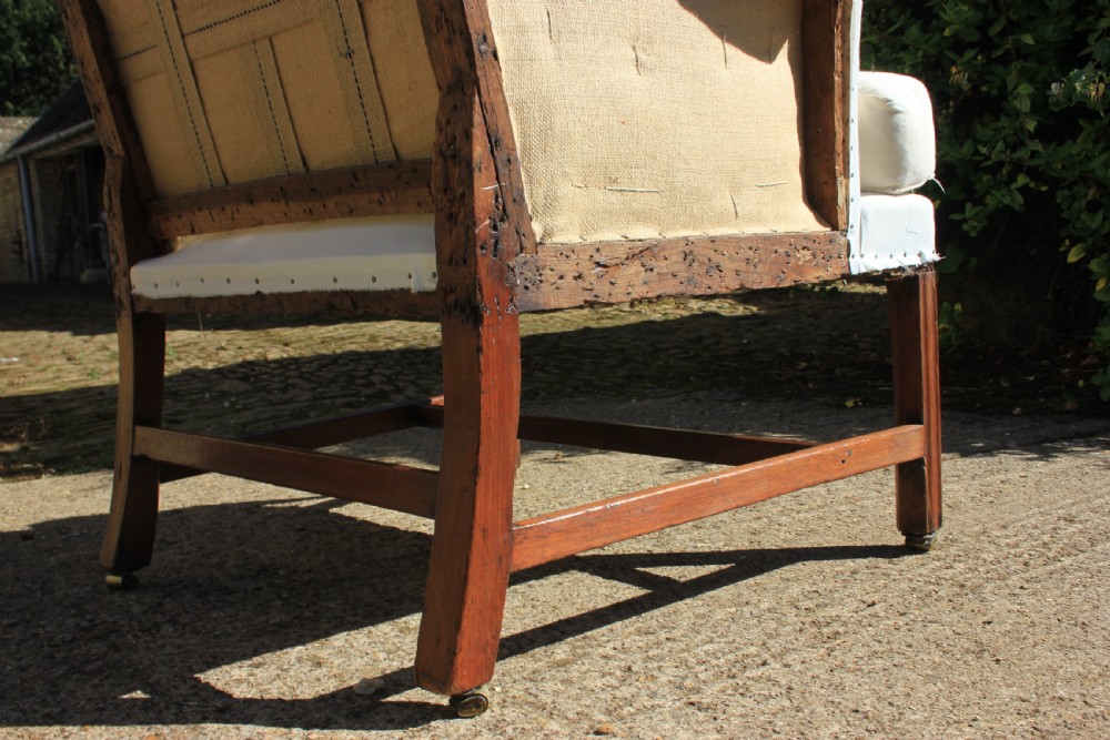 An 18th-century wingback armchair upholstered in off-white calico lining with dark mahogany legs, an H-stretcher frame, and brass castors. View VI