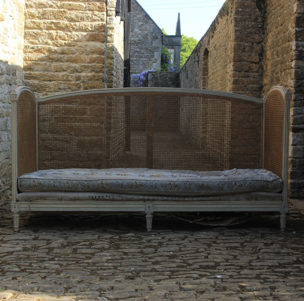 A French Louis XVI style painted wood daybed sofa featuring hand-woven cane panels along the arched back and sides, resting on fluted tapered legs with an upholstered cushion. View VI
