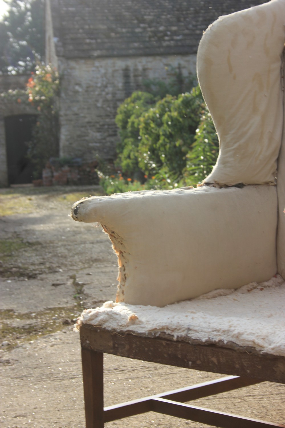 An early 18th-century wingback armchair featuring a dark timber frame, prominent side wings, and worn beige calico upholstery. View V