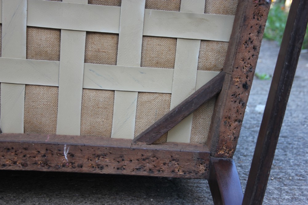 An early 18th-century wingback armchair featuring a dark timber frame, prominent side wings, and worn beige calico upholstery. View X