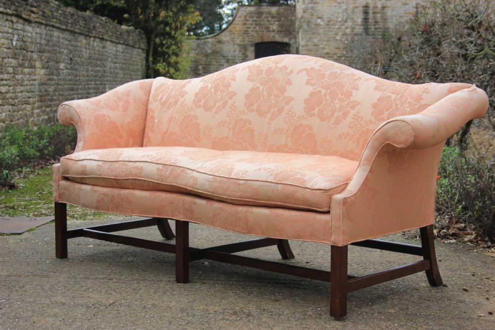 A peach damask upholstered camel back sofa featuring a serpentine seat cushion, out-scrolled arms, and square mahogany legs connected by stretchers.