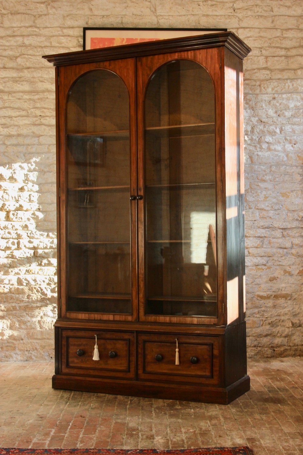 A 19th-century mahogany glazed bookcase featuring two arched glass doors, internal wooden shelving, and two lower drawers with turned wooden knobs.