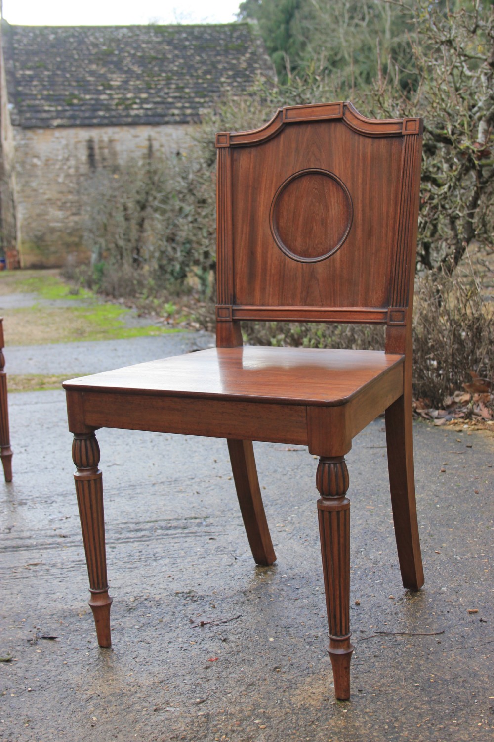 A pair of mahogany hall chairs featuring carved backrests with central circular mouldings, solid timber seats, and fluted turned front legs. View III