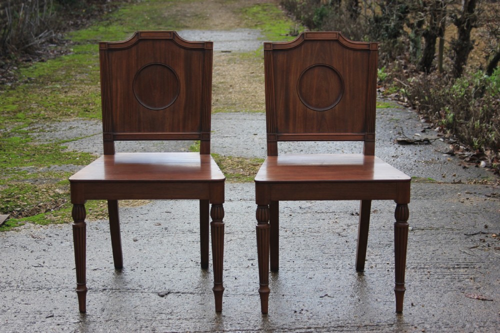 A pair of mahogany hall chairs featuring carved backrests with central circular mouldings, solid timber seats, and fluted turned front legs. View VII