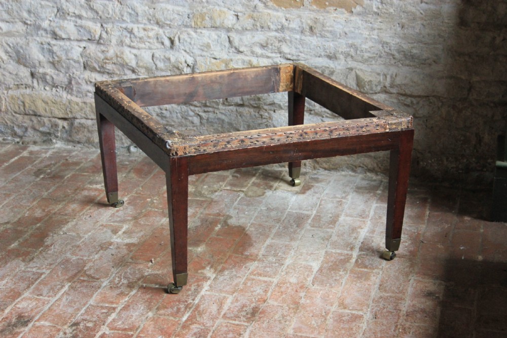 A square mahogany footstool frame with tapered legs and brass castors, showing exposed tacking rails where former upholstery was removed.