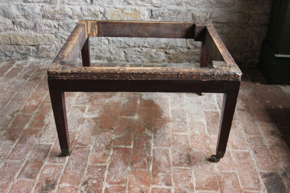 A square mahogany footstool frame with tapered legs and brass castors, showing exposed tacking rails where former upholstery was removed. View V