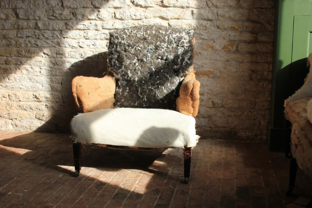 An antique Howard style armchair with exposed calico and horsehair padding, scrolled arms, and turned wooden front legs on brass castors. View IV