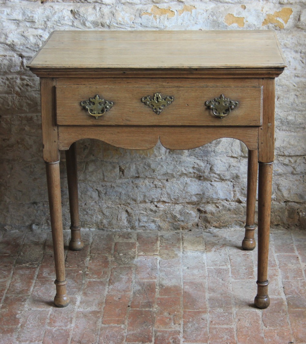 An 18th century pale oak lowboy featuring a single drawer with brass handles, a scalloped apron, and slender turned legs.