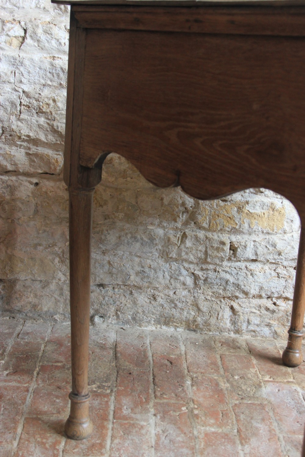 An 18th century pale oak lowboy featuring a single drawer with brass handles, a scalloped apron, and slender turned legs. View V