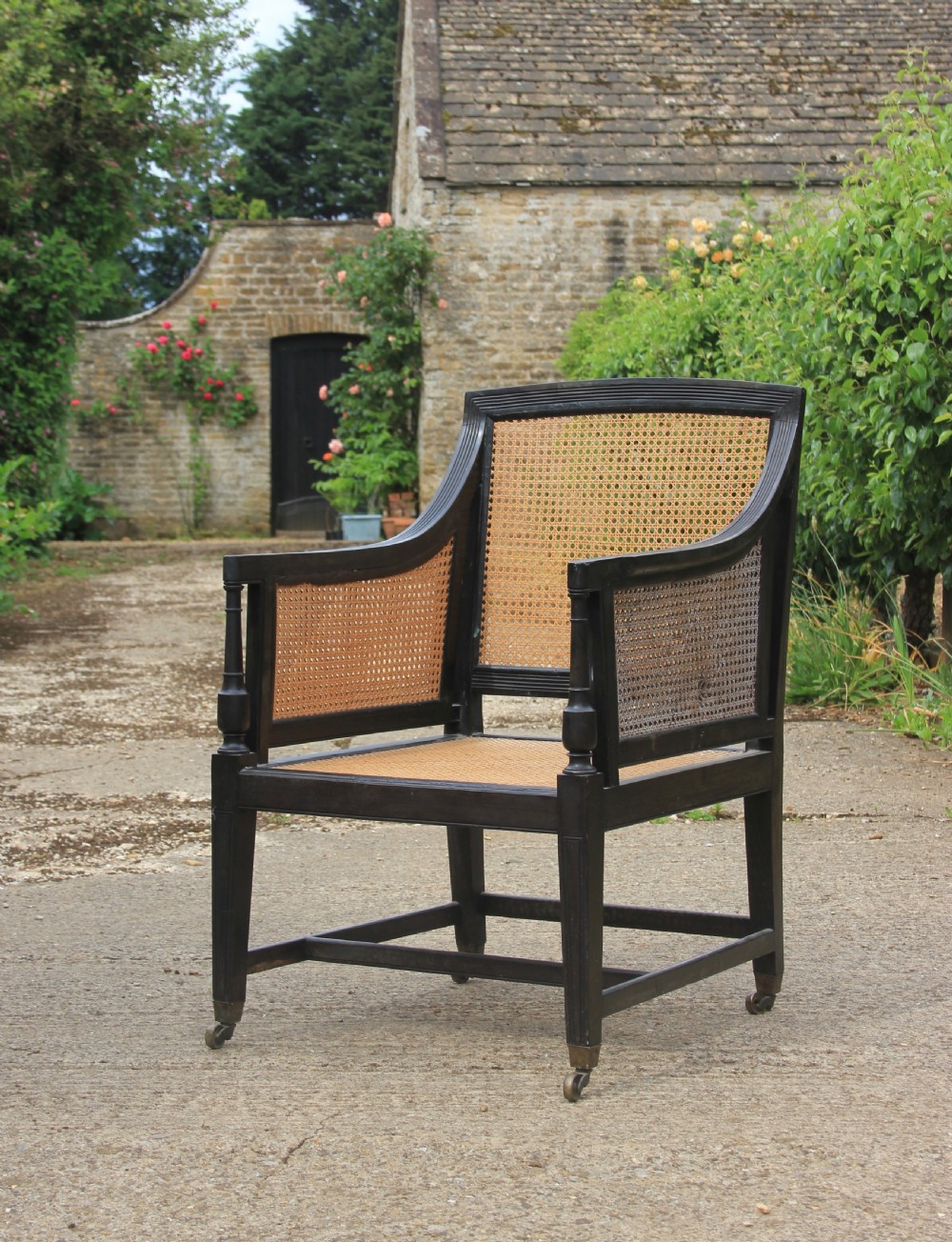An Anglo-Indian style dark ebony bergere armchair featuring woven cane panelling on the backrest, sides, and seat, resting on straight legs with brass castors. View II