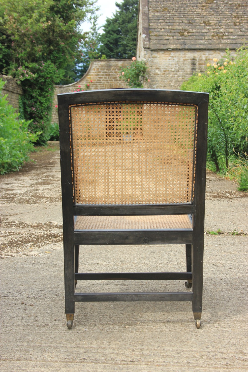 An Anglo-Indian style dark ebony bergere armchair featuring woven cane panelling on the backrest, sides, and seat, resting on straight legs with brass castors. View V