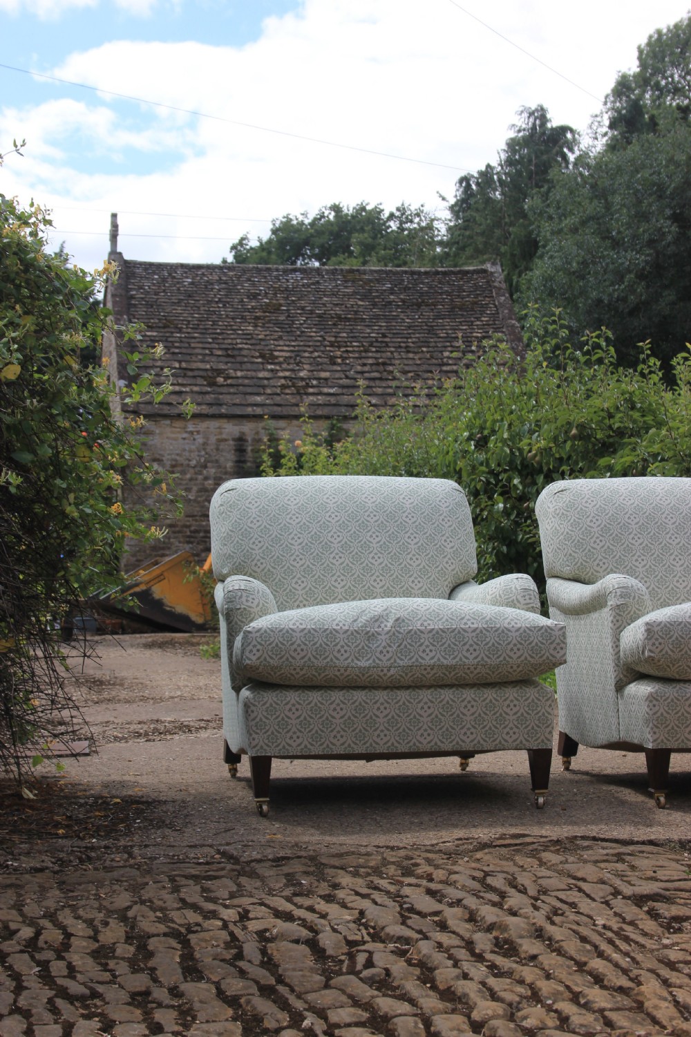 A pair of deep-seated armchairs upholstered in a sage green and off-white patterned fabric with turned front legs on brass castors. View II