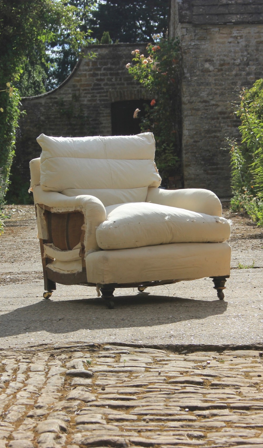 A deep-seated Howard style armchair covered in cream calico under-upholstery, supported by turned wooden front legs with brass castors.