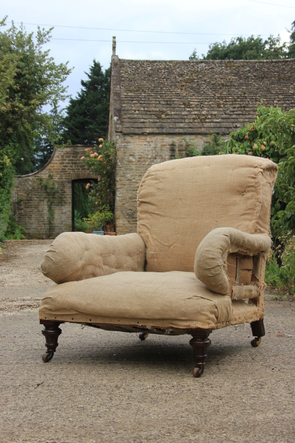 A Howard style armchair stripped down to its neutral hessian under-upholstery, featuring turned front wood legs set on brass castors.