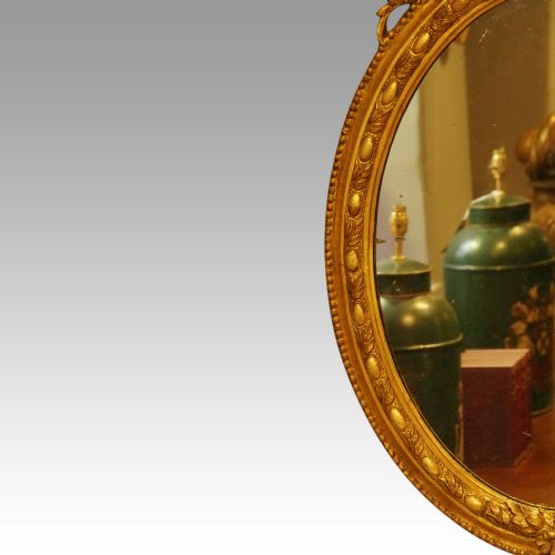 fig 1. An oval giltwood wall mirror featuring a carved pediment crest with scrolling details and a beaded rim.