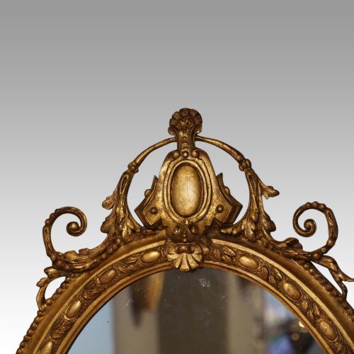 fig 2. An oval giltwood wall mirror featuring a carved pediment crest with scrolling details and a beaded rim.