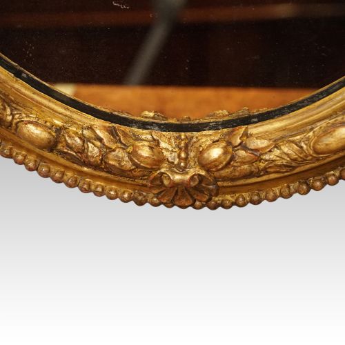 fig 3. An oval giltwood wall mirror featuring a carved pediment crest with scrolling details and a beaded rim.