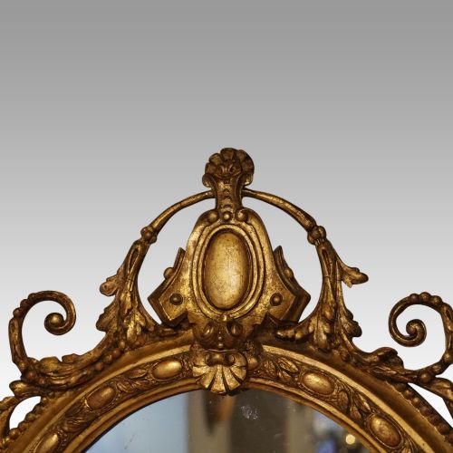fig 4. An oval giltwood wall mirror featuring a carved pediment crest with scrolling details and a beaded rim.