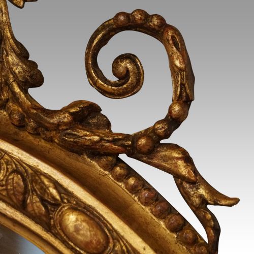 fig 5. An oval giltwood wall mirror featuring a carved pediment crest with scrolling details and a beaded rim.