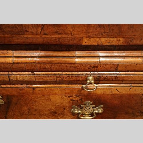 fig 7. A George II walnut tallboy featuring stacked drawers with decorative brass handles, bracket feet, and a sunburst motif on the bottom drawer.