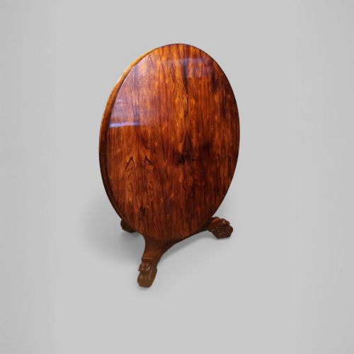 fig 1. A circular antique rosewood dining table with a highly figured timber top shown upright above a carved tri-form pedestal base.