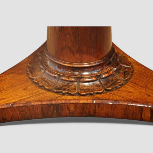 fig 2. A circular antique rosewood dining table with a highly figured timber top shown upright above a carved tri-form pedestal base.