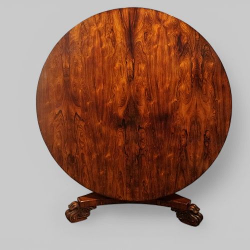 fig 7. A circular antique rosewood dining table with a highly figured timber top shown upright above a carved tri-form pedestal base.