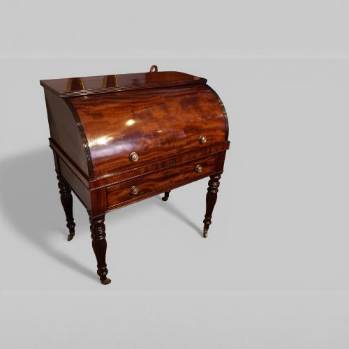 fig 1. An early nineteenth-century mahogany cylinder desk featuring a curved roll-front cover, brass knobs, a long frieze drawer, and turned reeded legs with brass castors.