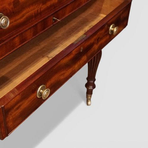 fig 4. An early nineteenth-century mahogany cylinder desk featuring a curved roll-front cover, brass knobs, a long frieze drawer, and turned reeded legs with brass castors.