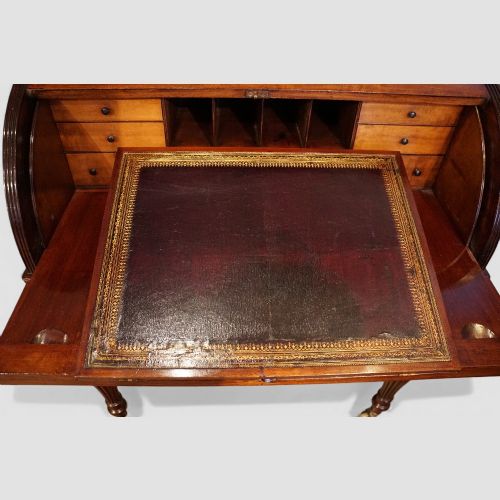 fig 7. An early nineteenth-century mahogany cylinder desk featuring a curved roll-front cover, brass knobs, a long frieze drawer, and turned reeded legs with brass castors.