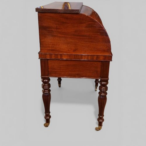 fig 9. An early nineteenth-century mahogany cylinder desk featuring a curved roll-front cover, brass knobs, a long frieze drawer, and turned reeded legs with brass castors.